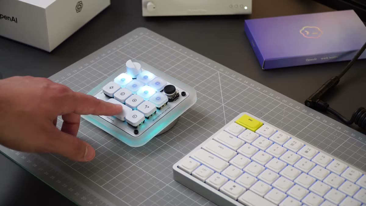 The Codex Micro sitting next to a white low-profile keyboard on a cutting mat. Behind it are an OpenAI-branded white box and a purple OpenAI x Work Louder box. A hand is pressing one of the keys.