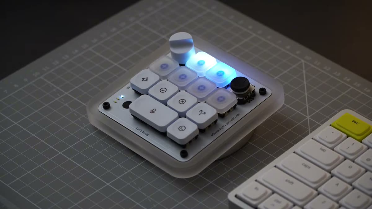 The Codex Micro macro pad on a cutting mat: a square white deck with a translucent joystick, a black rotary dial, and a grid of white keys marked with icons. Several frosted keys glow blue. The base is engraved 'You can just build things.'